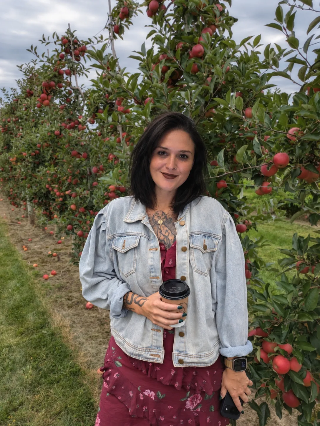 Ashley smiling in an apple orchard, wearing a denim jacket over a burgundy floral dress and holding a coffee, with rows of red apples behind her