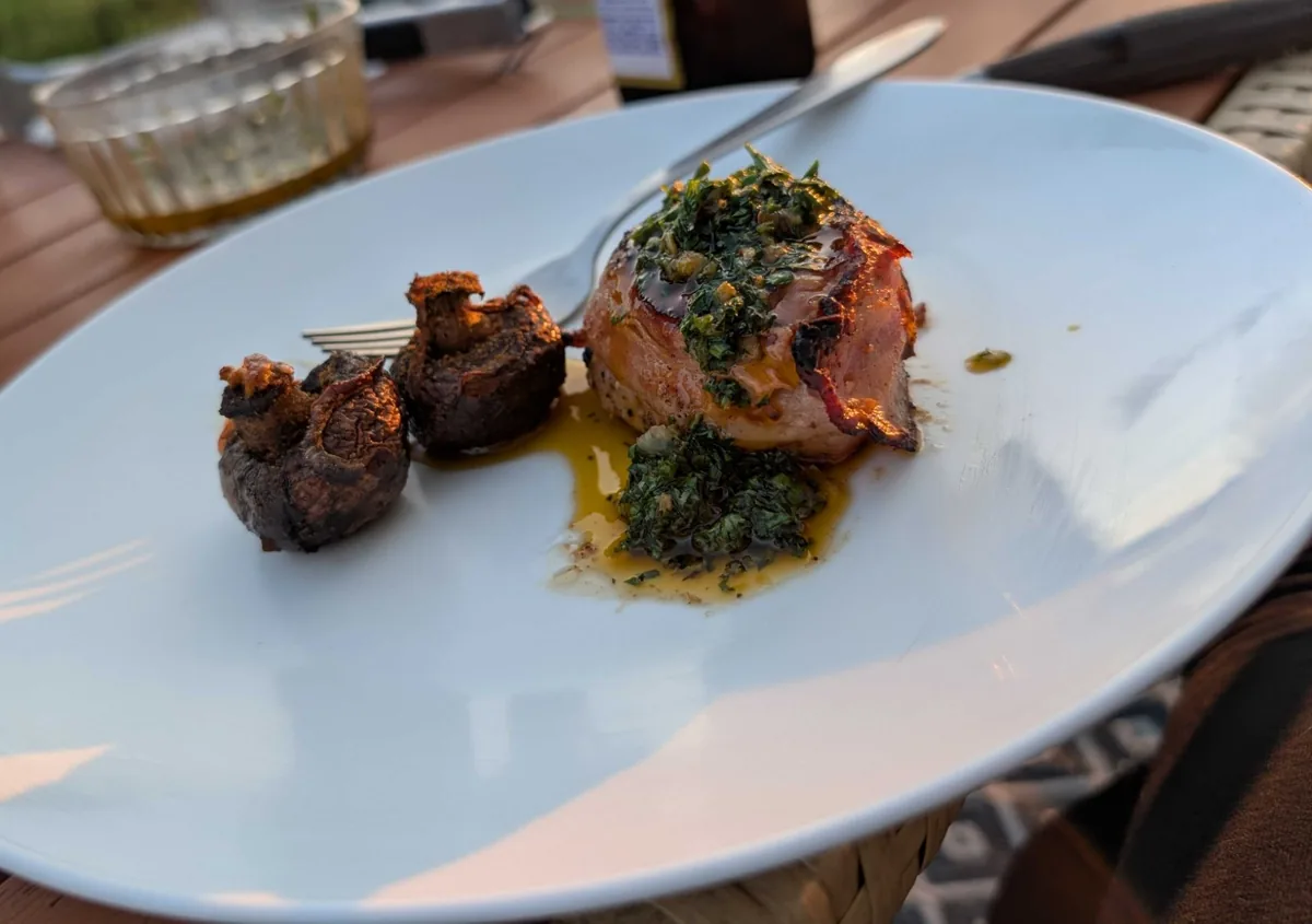 A bacon-wrapped filet mignon topped with chimichurri next to two smoked baby bellas, on a white plate outdoors in evening sun