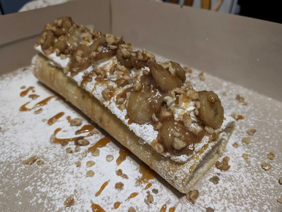 A sponge roll dusted in powdered sugar, topped with caramel bananas and chopped candied pecans, caramel drizzled across the board