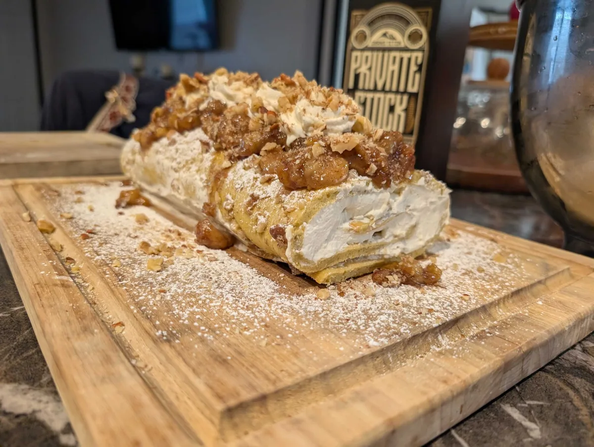 The bananas foster roll from the cut end, showing the cream spiral under caramel bananas, piped cream and chopped candied pecans on a powdered-sugar board