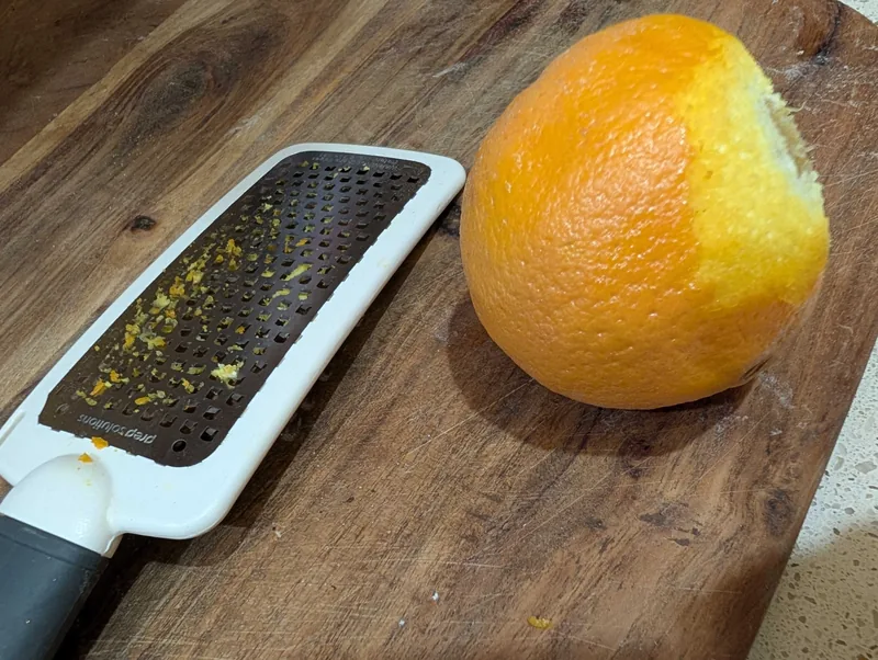 A half-zested orange on a wooden board next to a fine grater flecked with zest
