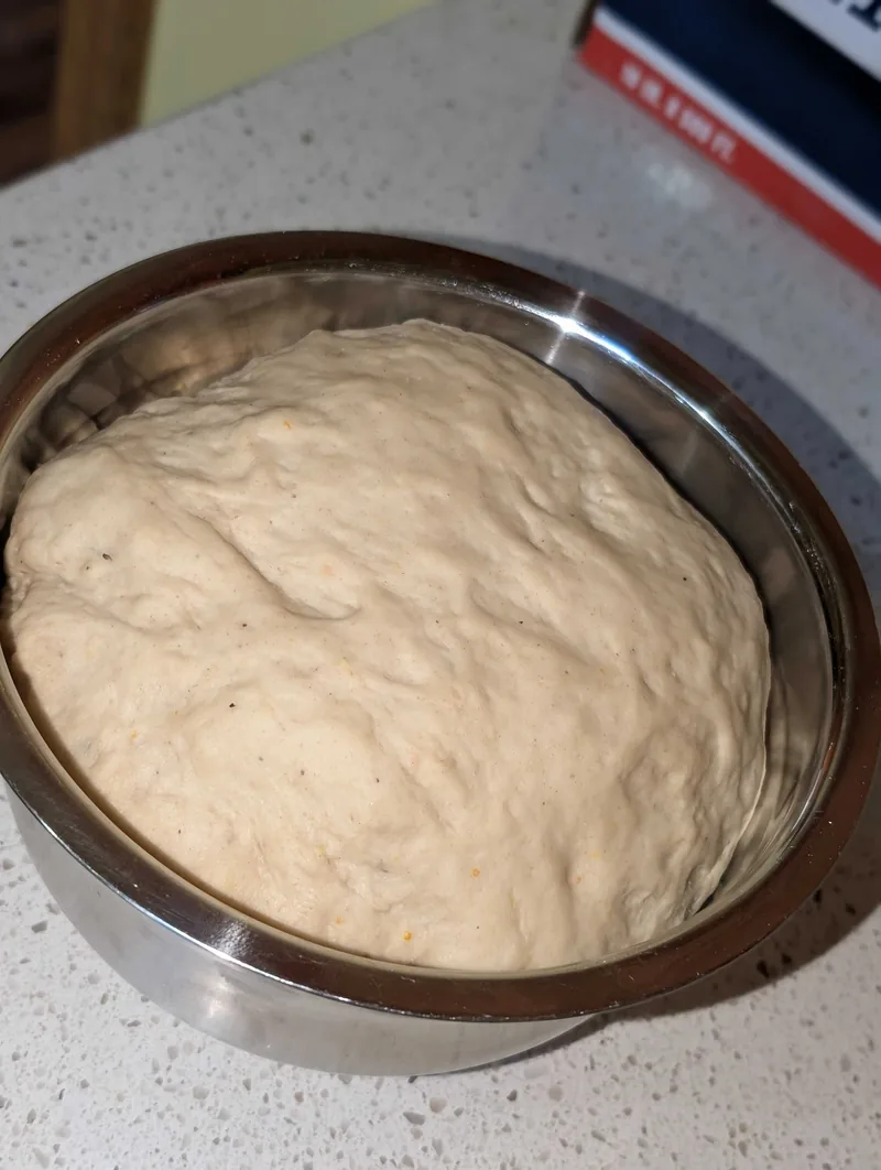 The dough risen and domed in a steel bowl, flecked with orange zest and black pepper