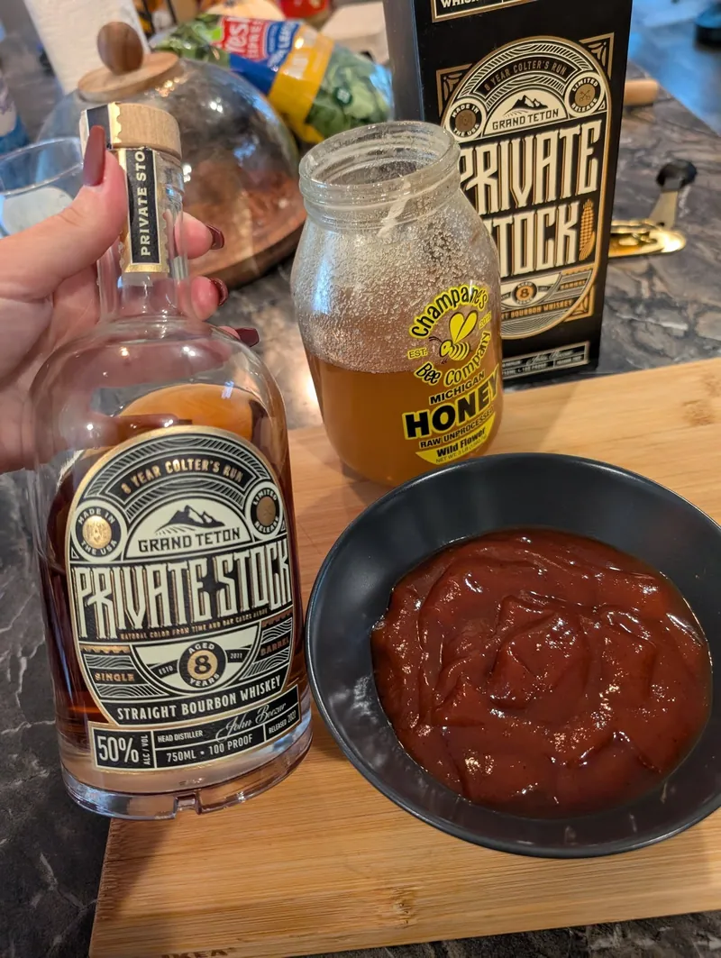 A bottle of Grand Teton Private Stock bourbon, a jar of Michigan wildflower honey, and a black bowl of finished barbecue glaze