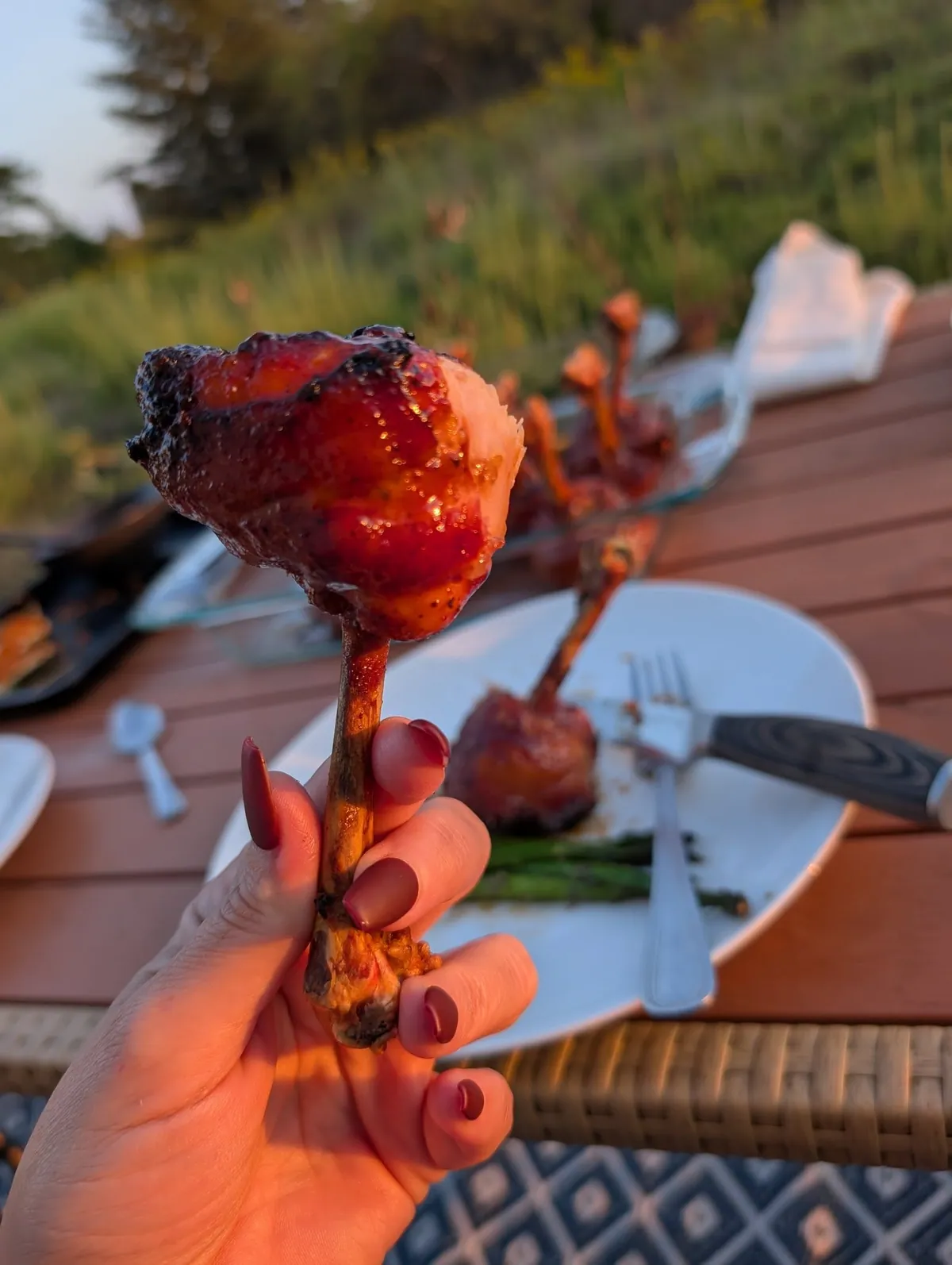 A hand holding a finished chicken lollipop by the bone, lacquered and charred at the edges, in evening light