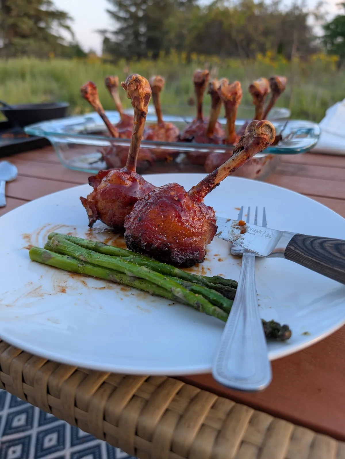 Two glazed chicken lollipops on a white plate with charred asparagus, on a deck table at sunset with the tray behind