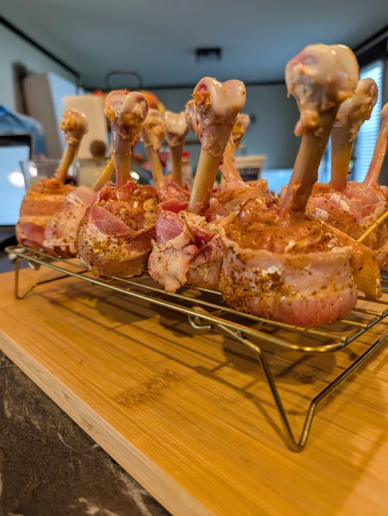 A rack of raw bacon-wrapped chicken lollipops on a bamboo board, bones frenched clean and pointing up
