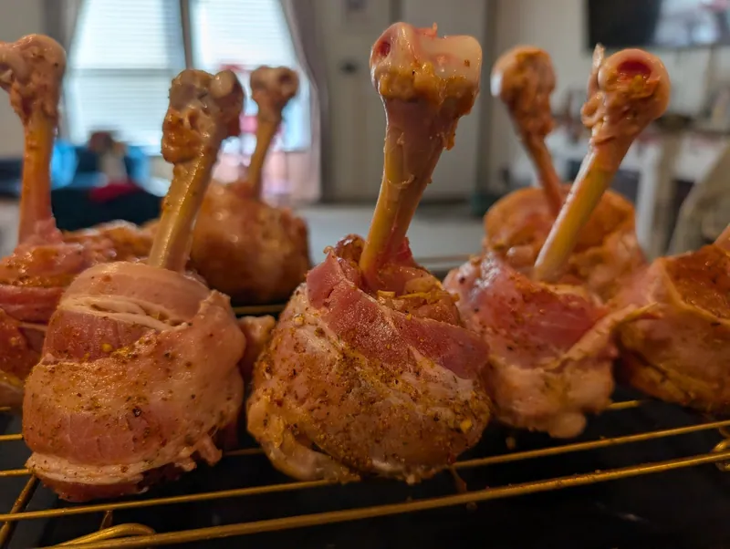 Raw bacon-wrapped chicken lollipops standing on a wire rack, seasoned and ready for the smoker