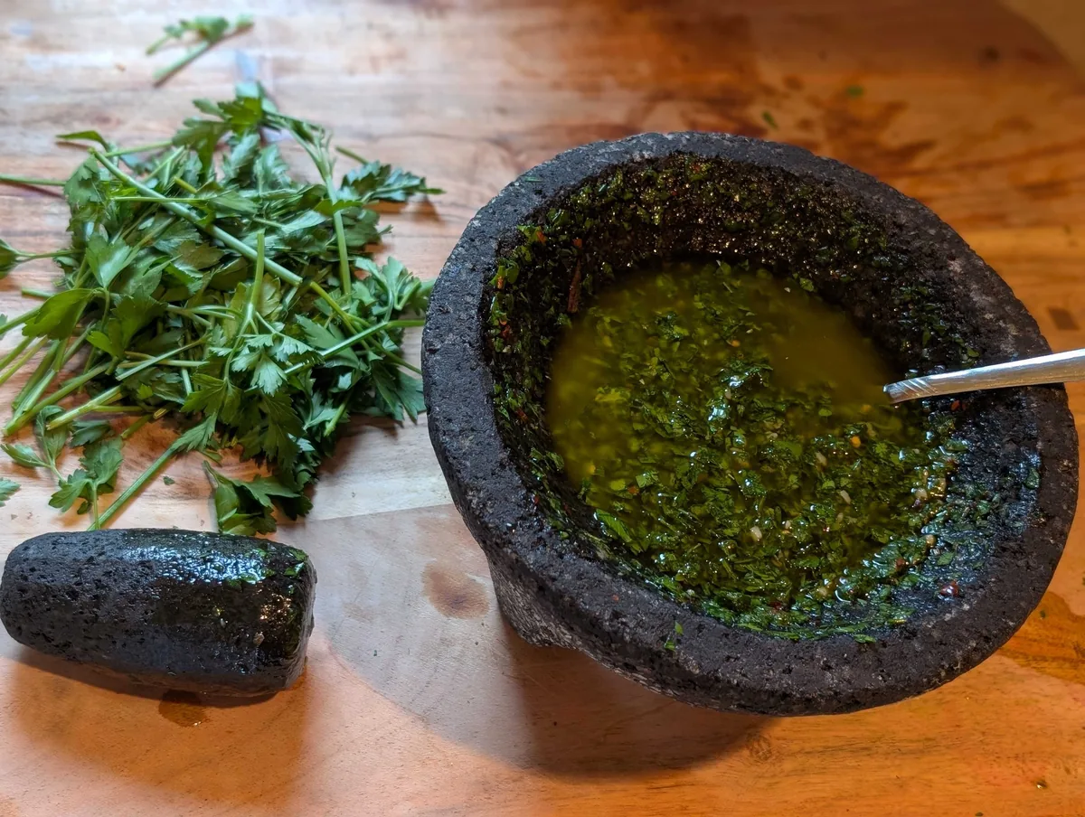 Chimichurri in a black stone molcajete with a spoon, a pile of fresh parsley and the pestle on a wooden board