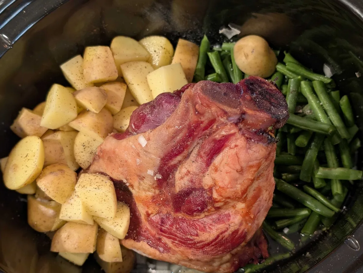 A smoked ham hock in a black crockpot between seasoned little potatoes and cut green beans, seen from above