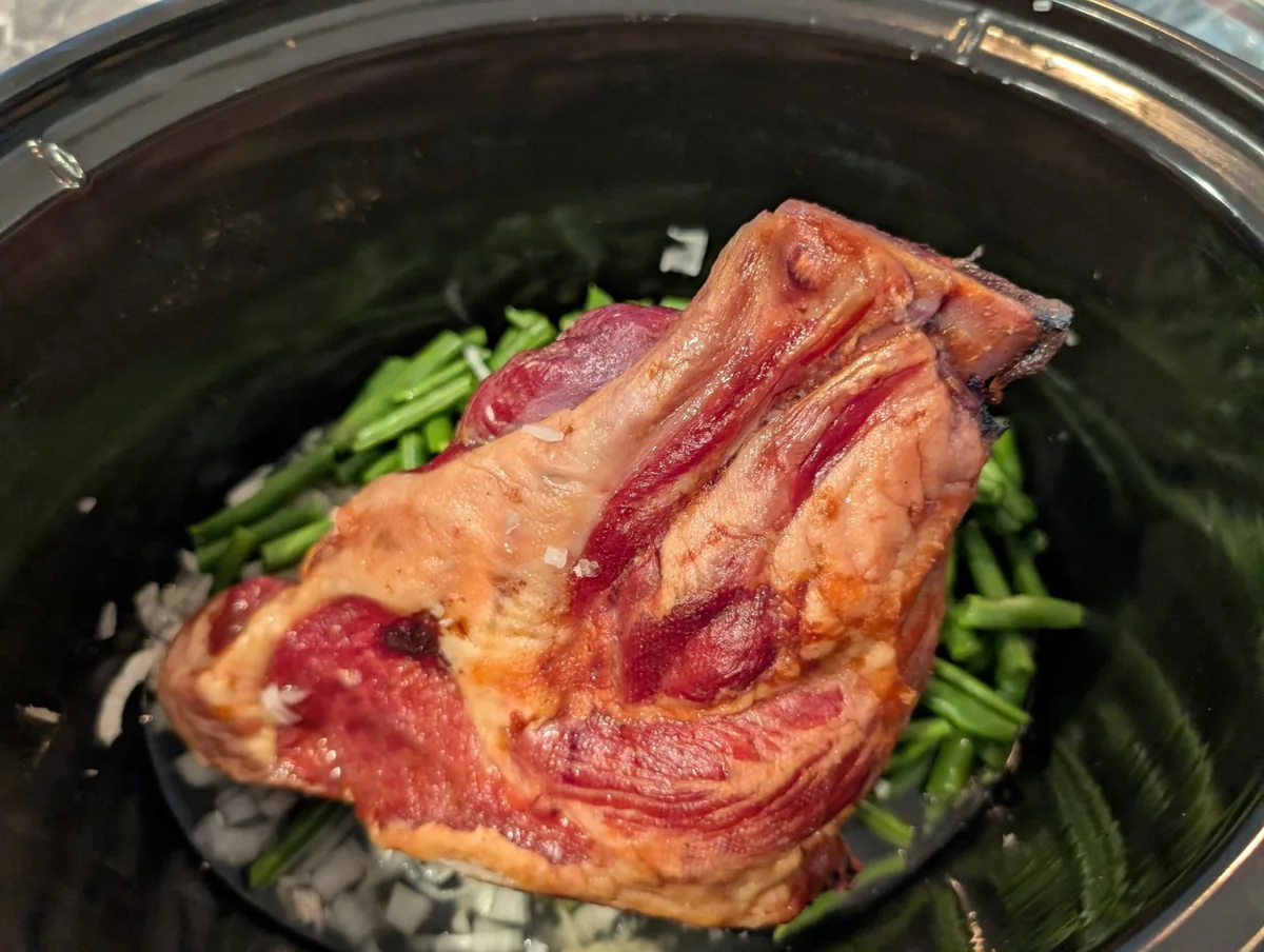 A glossy smoked ham hock resting on green beans and diced onion in the crockpot, before the potatoes go in