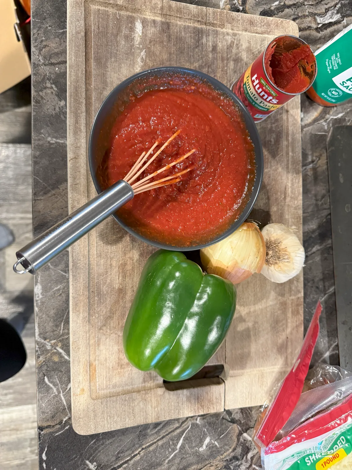 A pan of tomato sauce with a whisk on a cutting board, beside a green bell pepper, a yellow onion and a head of garlic