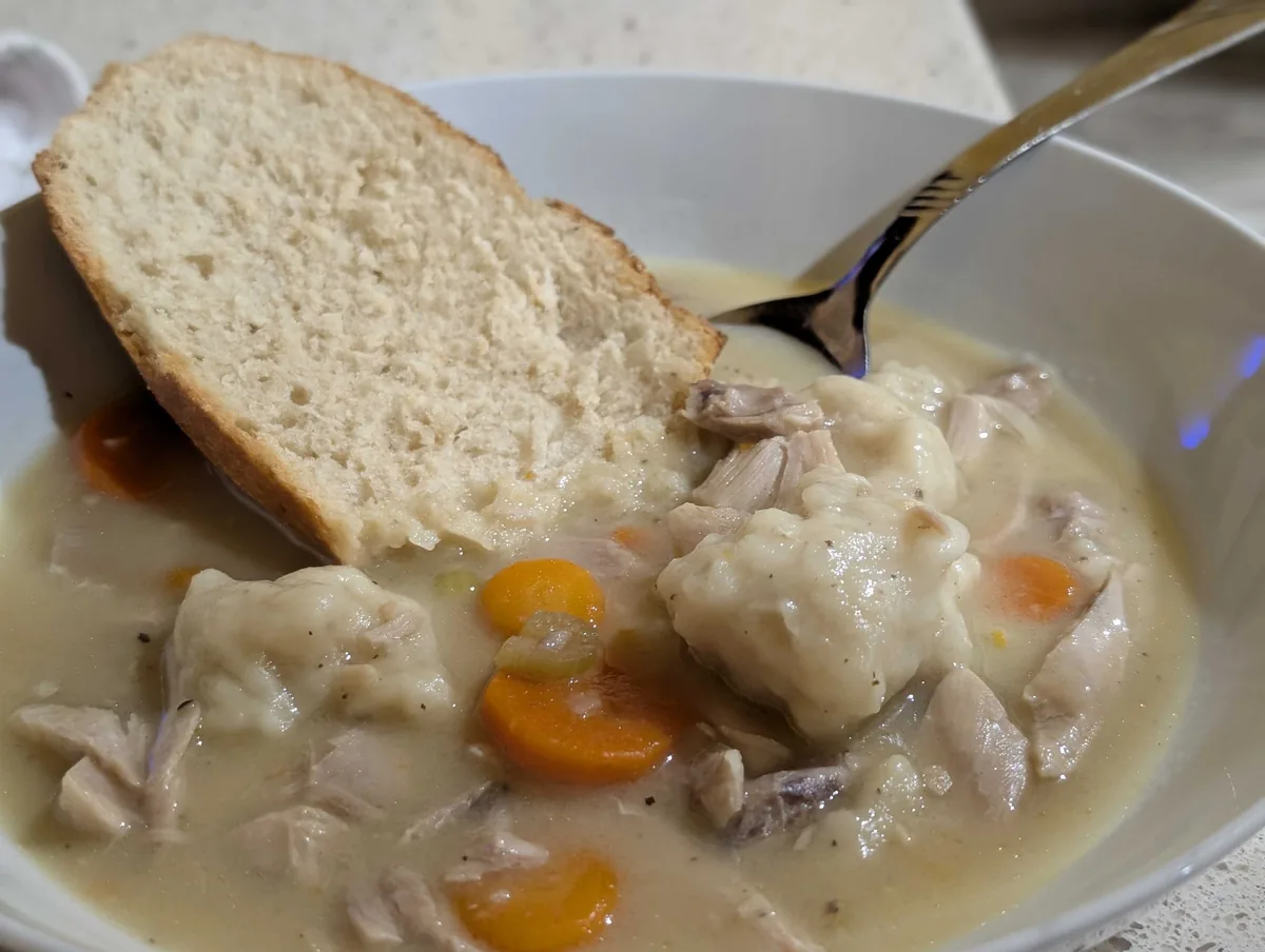 A bowl of creamy chicken and dumplings with carrots and pulled chicken, a thick slice of the Blue Moon bread dipped into the gravy and a spoon resting on the rim