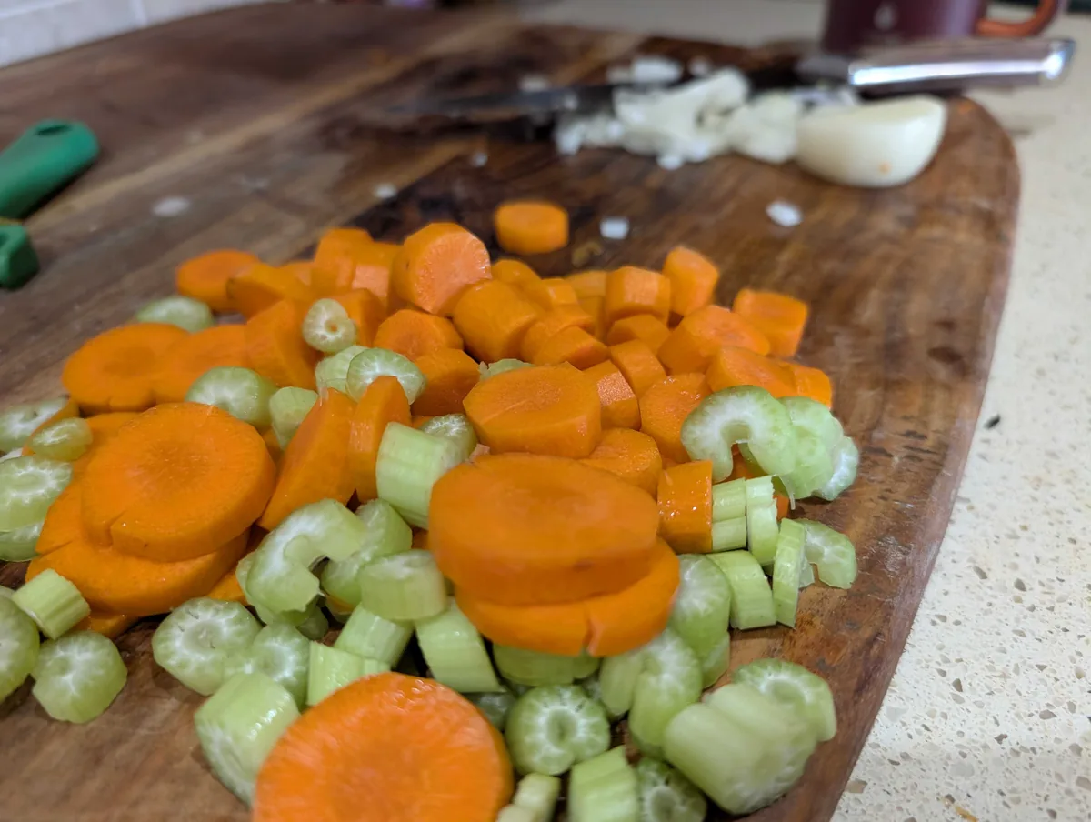 Sliced carrots and celery piled on a wooden board, with chopped onion and garlic behind them