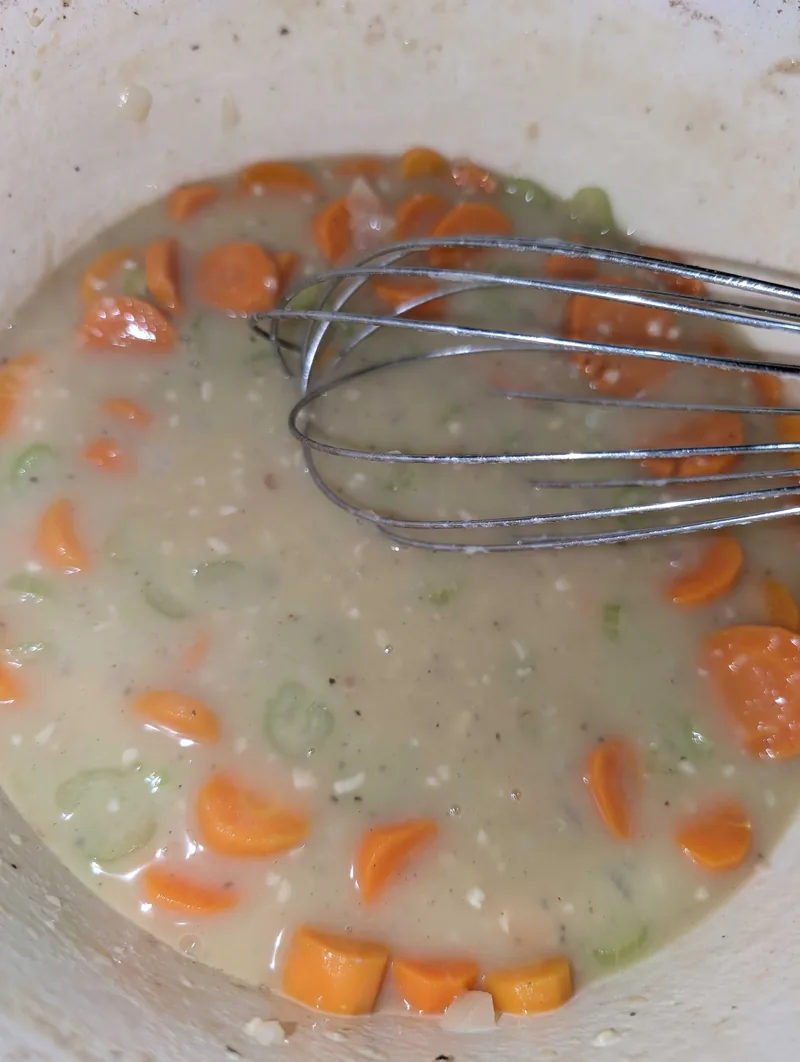 A whisk in the creamy gravy in a white Dutch oven, with sliced carrots and celery