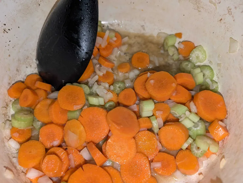 Carrots, celery and onion softening in browned butter in a white Dutch oven, a black spoon resting in the pot