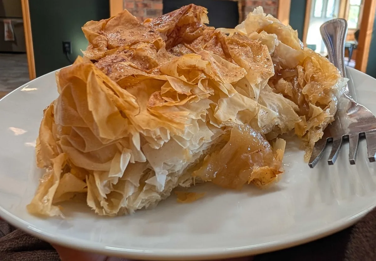 A slice of Gascon apple pie on a white plate, shattered filo layers over soft bourbon apples, a fork beside it