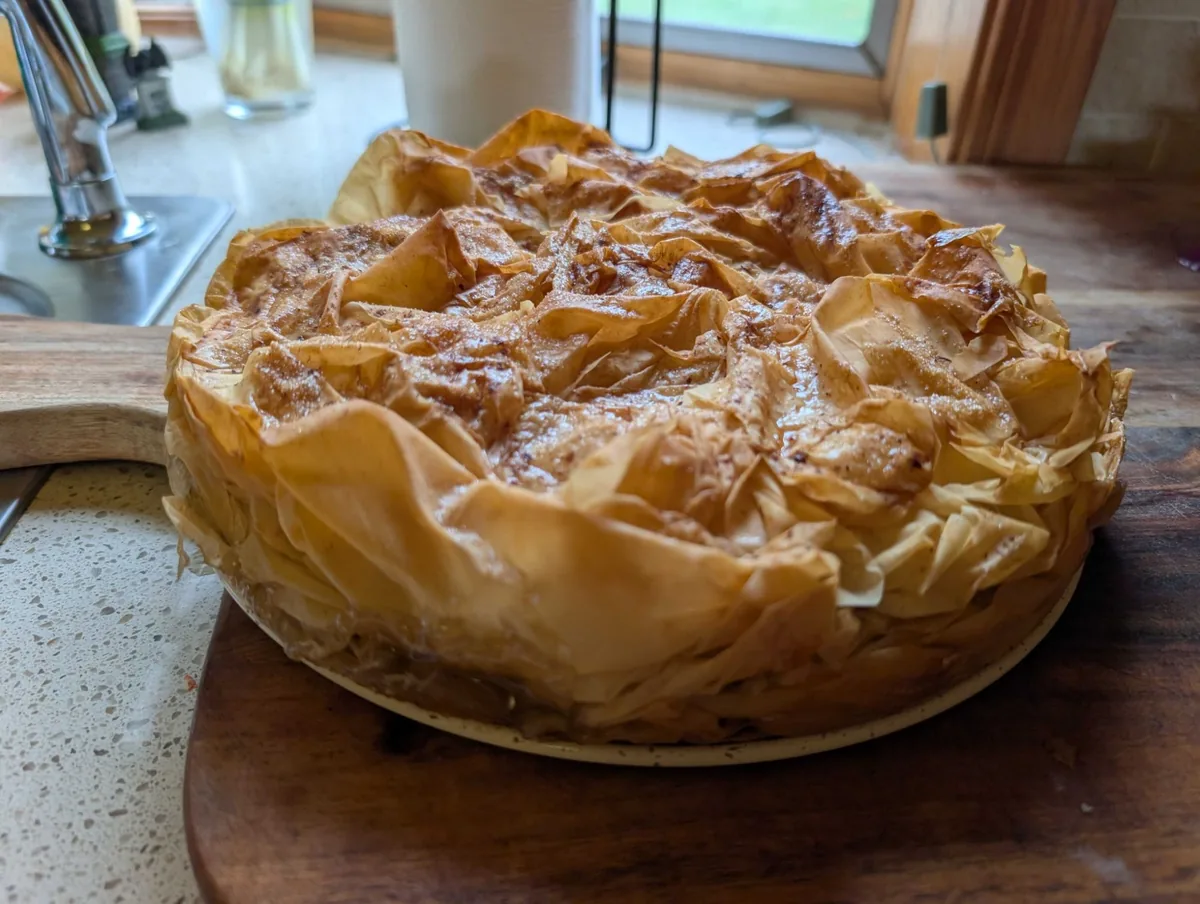 A whole Gascon apple pie with a tall crown of scrunched, golden filo, on a wooden board by the kitchen sink