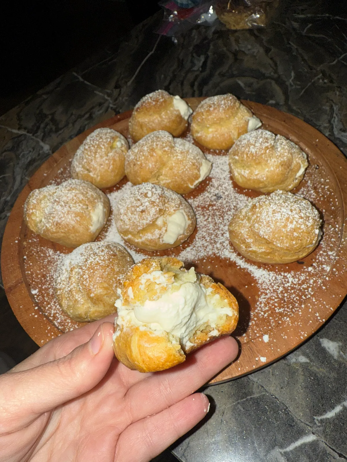A hand holding a cream puff broken open to show the vanilla cream, over a board of powdered-sugar-dusted puffs