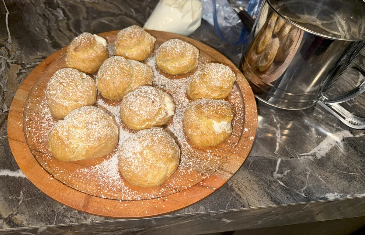 A round wooden board of golden cream puffs dusted with powdered sugar, a sifter beside it on a dark marble counter