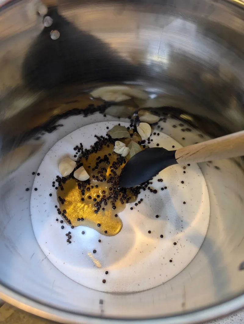Looking down into a steel pot at salt, black peppercorns, smashed garlic and bay leaves for the brine