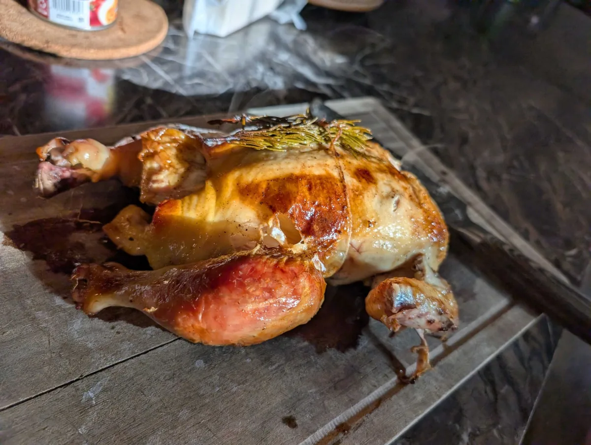 A whole roast chicken on a board, skin golden and glossy with a sprig of rosemary tied across the breast