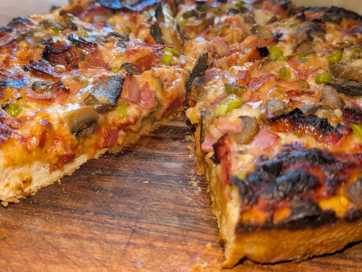 A cut edge of the pizza showing the thick, airy IPA crust under the toppings and the crisp, darkened cheese edge