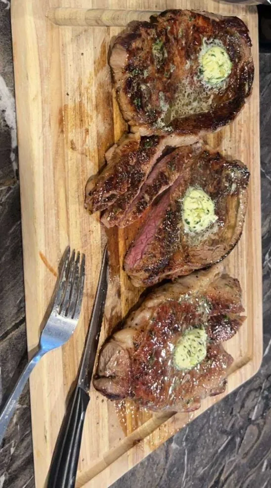 Three seared steaks on a wooden cutting board, each topped with a melting coin of green herb butter, one sliced to show a pink medium center