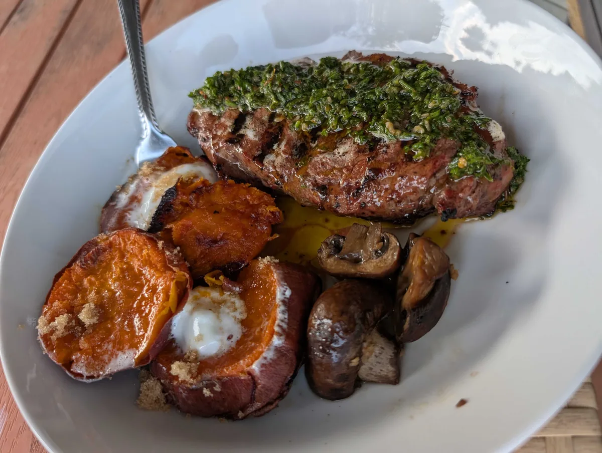 A grilled steak topped with bright green chimichurri in a white bowl, with roasted sweet potato rounds and mushrooms