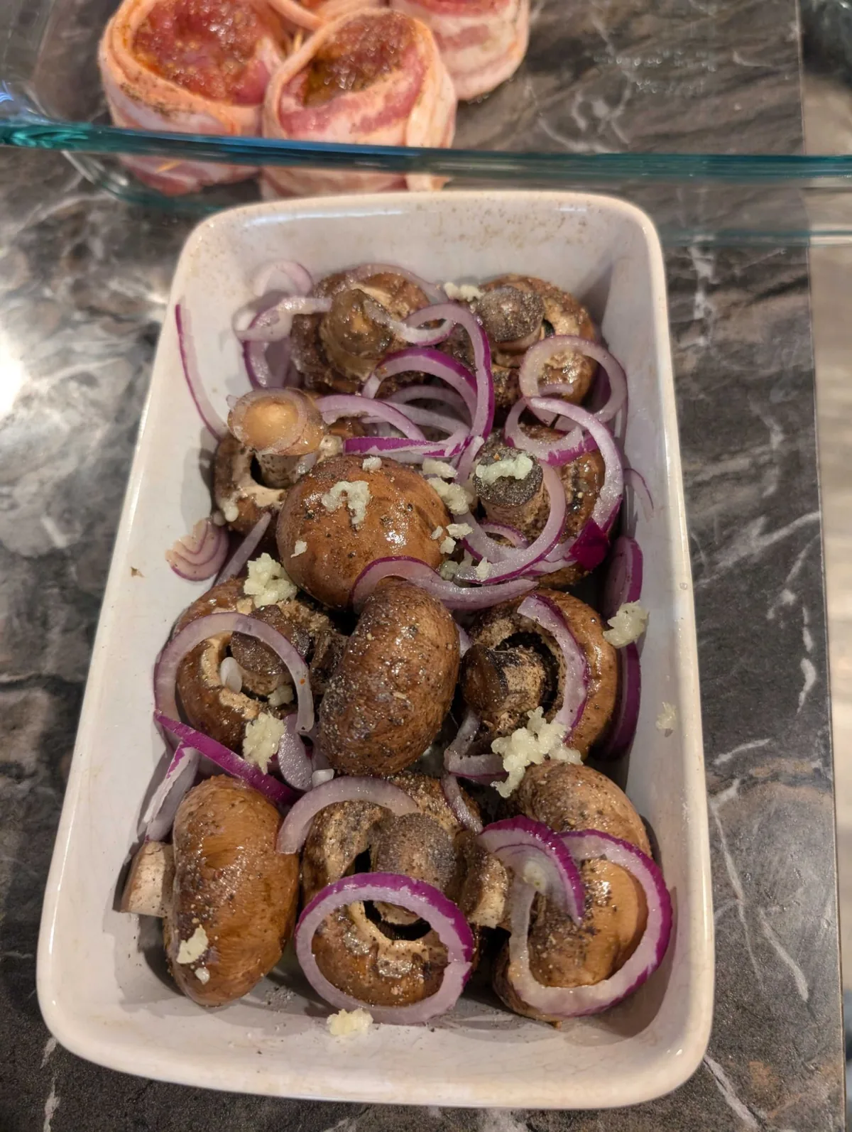 Whole baby bellas with sliced onion, chopped garlic and cracked pepper packed into a dish, bacon-wrapped steaks waiting behind