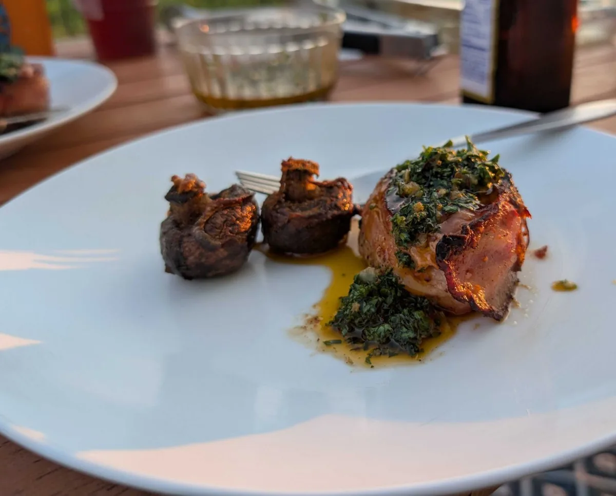 Two smoked baby bella mushrooms beside a bacon-wrapped steak topped with chimichurri, on a white plate outdoors at sunset