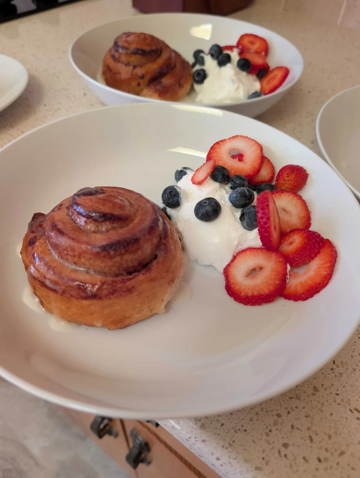 A golden cinnamon roll in a wide white bowl beside a scoop of yogurt with blueberries and sliced strawberries