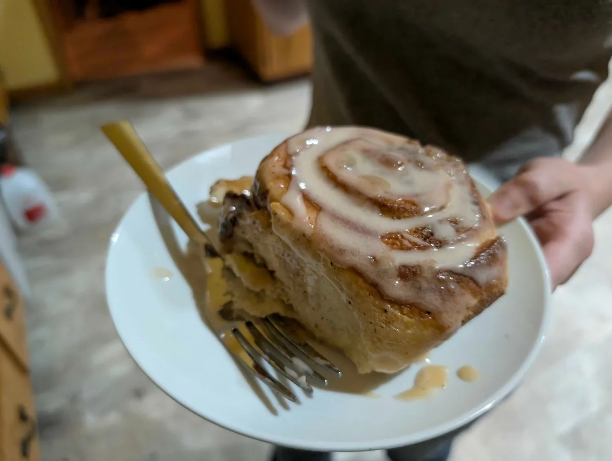 An iced cinnamon roll on a small white plate with a gold fork, icing running down its sides