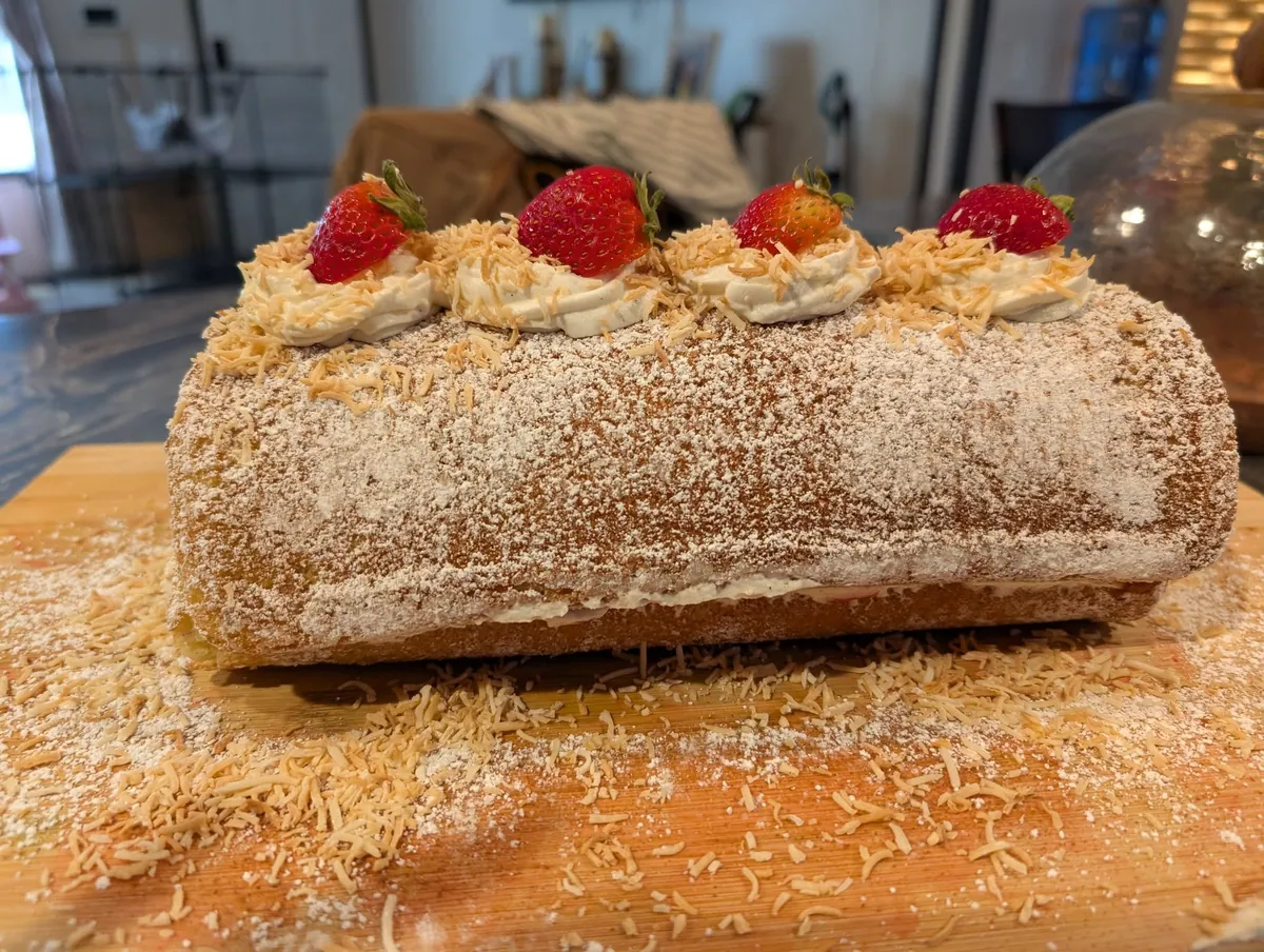 A sponge roll under a heavy dusting of powdered sugar and toasted coconut, topped with four cream rosettes each crowned with half a strawberry