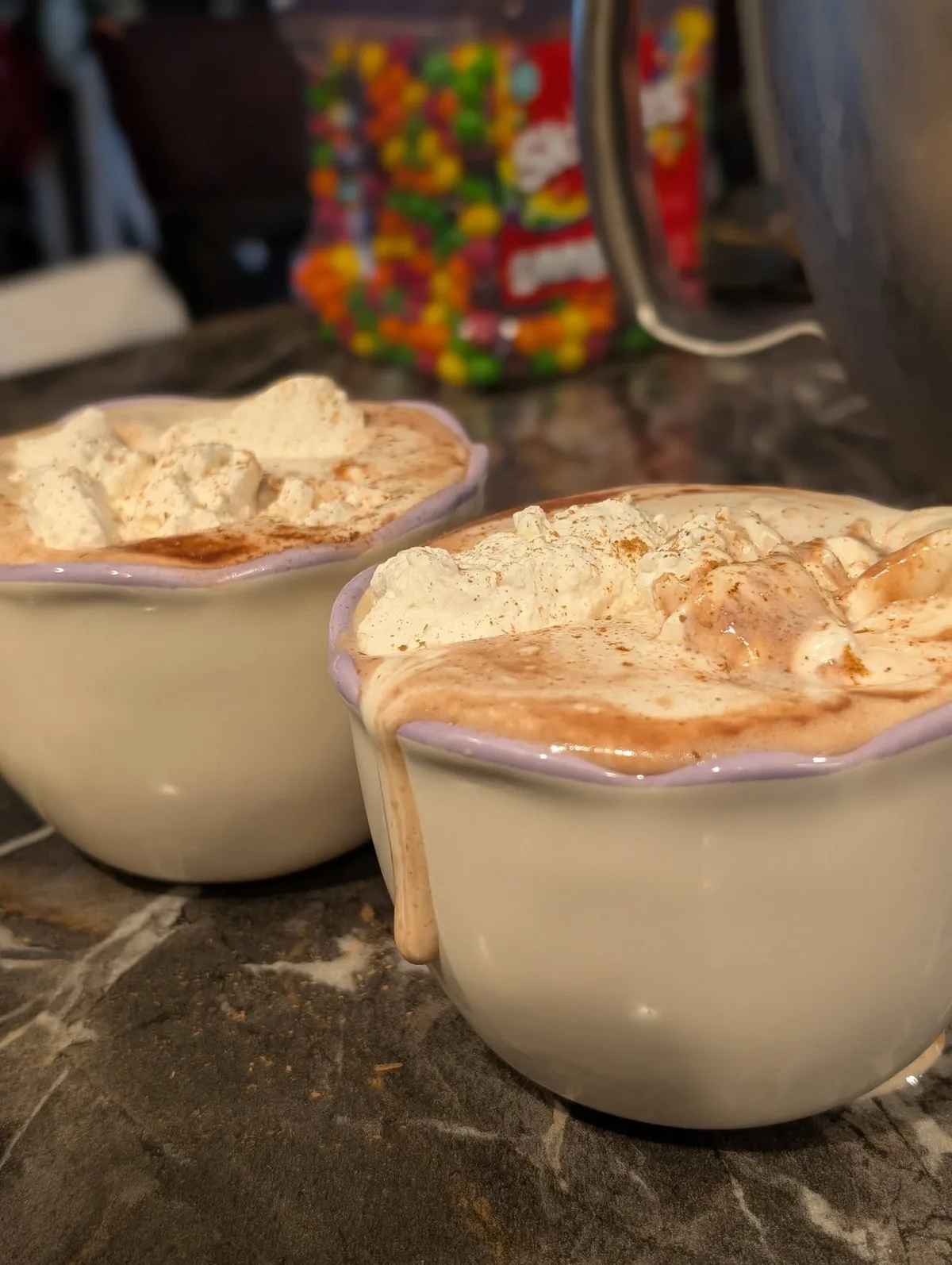 Two cream cups of hot chocolate, piled high with whipped cream, on a dark marble counter
